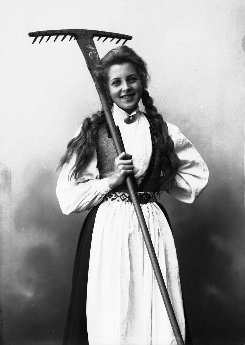 Solveig Lund, 3/4 dels studioportrett av kvinne i drakt, med flettet hår. Hun holder en rive i høyre arm, mens den venstre hviler mot hoften. Bakgrunnen er nøytral.