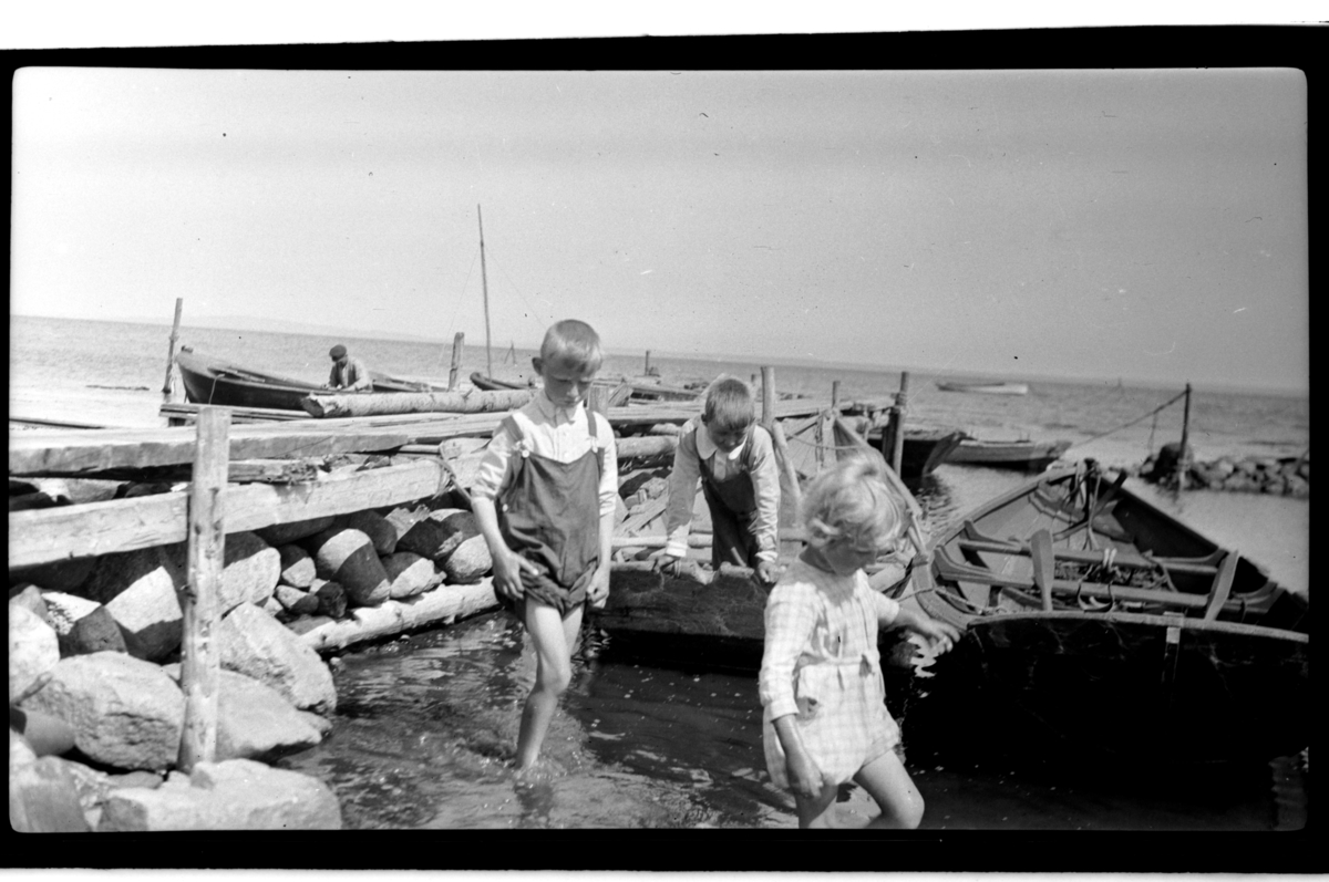 Rolf Sr. Sundt, Julius Sundt, en ukjent gutt og Rolf Sundt jr. vasser ved en brygge, flere robåter ligger fortøyd, Slagen. Fotografert 1925.
