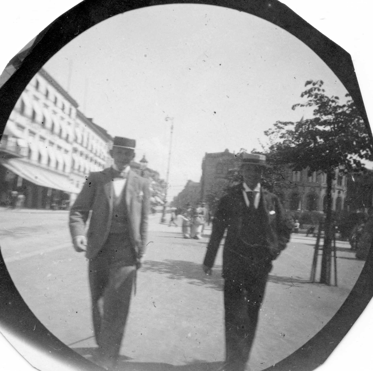 Fredrik Carl Mülertz Størmer, Stud. Niels Heitmann og Mohr spaserer langs Karl Johans gate, Oslo, med Stotinget i ryggen og Grand Hotel.på andre siden av gata.
