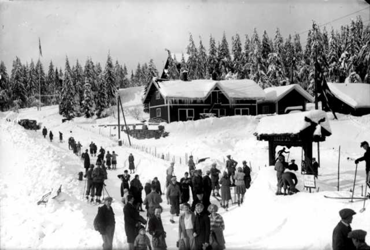 Hermann Christian Neupert, Frognerseteren, Nordmarka, Oslo. Vintermoltiv. Mennesker langs vei på bena og på ski. Hus i bakgrunnen,