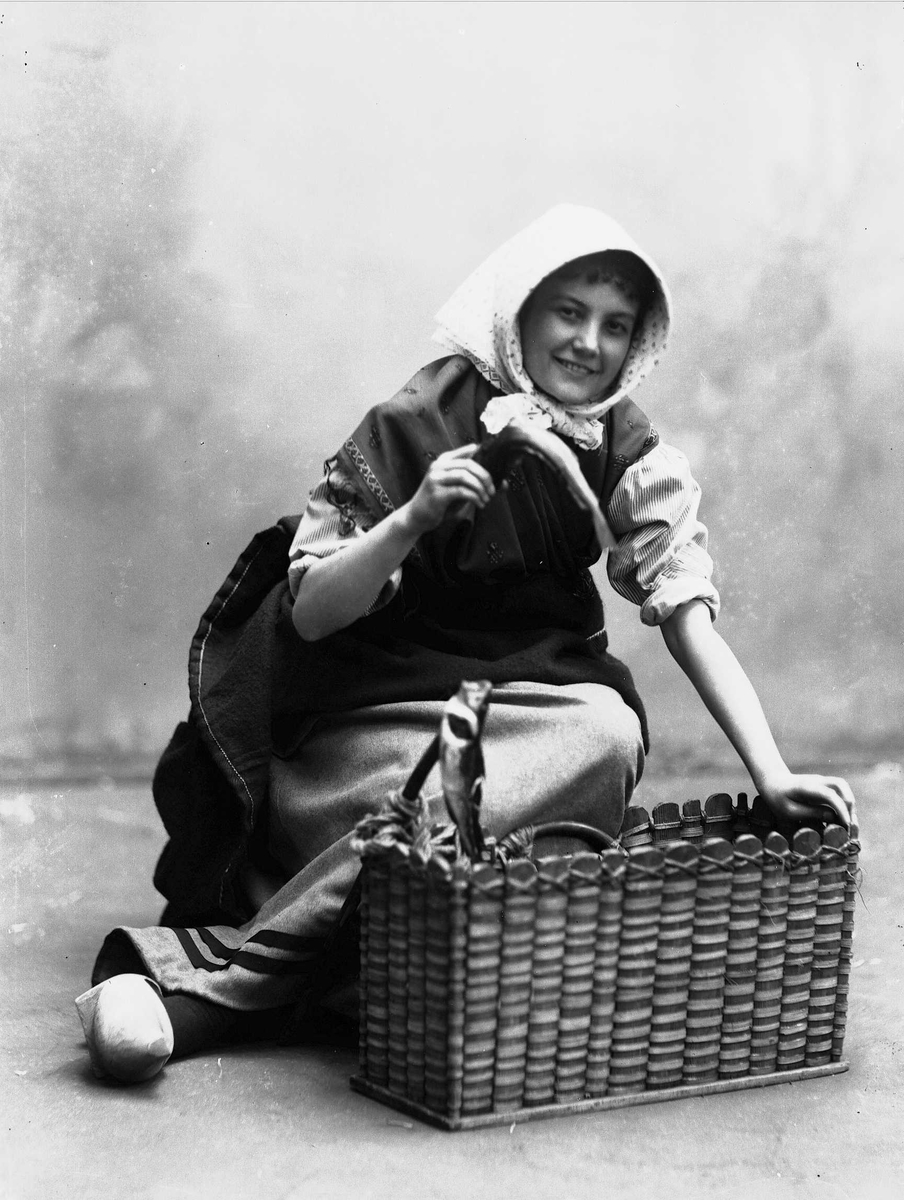 Solveig Lund, Studioportrett av kvinne i drakt med skaut på hodet. Hun sitter på gulvet foran en trekurv og holder en fisk i den ene hånden. Bakgrunnen er malt.