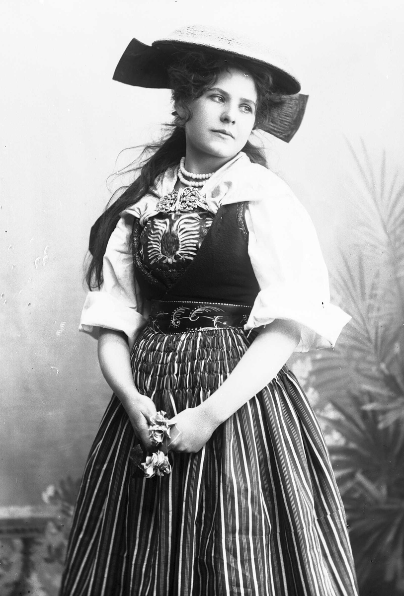 Solveig Lund, Kvinne i drakt og hatt, med blomst i hendene, plassert foran bakteppe med bregner og blomster.