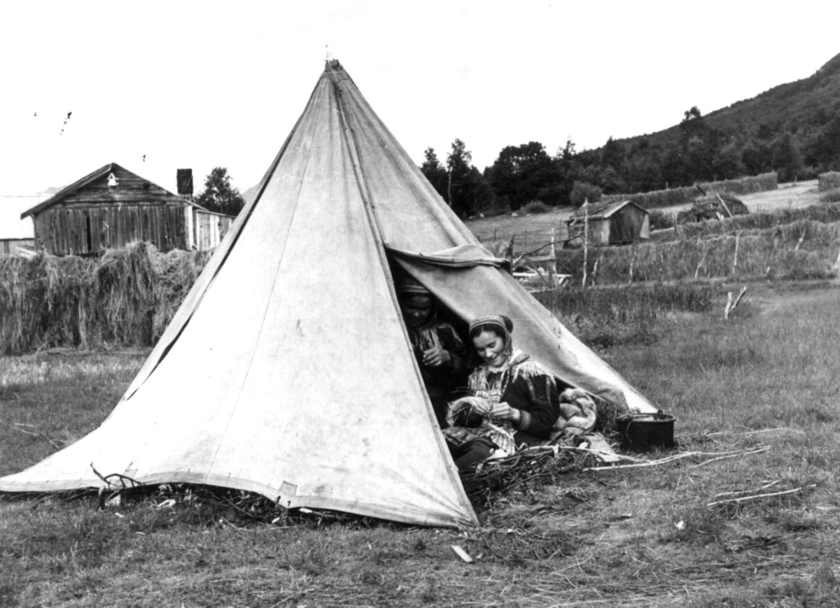 Anna Grostøl, To kvinner i åpningen til et sametelt, Hesjer og bygninger i bagrunnen. Olderdalen, Kåfjord, Troms 1947.