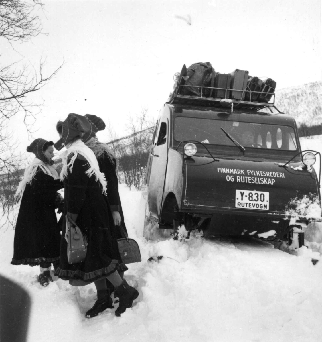 Virginia Raymond Davidson, Snowmobile. Snøbil med oppakking på taket sitter fast i snøen,ved siden av står tre samekvinner. Karasjok 1958. Denne beltevogna var registrert Y-830 med undertekst "Rutevogn". Den gikk i rutetrafikk hos Finnmark Fylkesrederi og Ruteselskap (FFR).