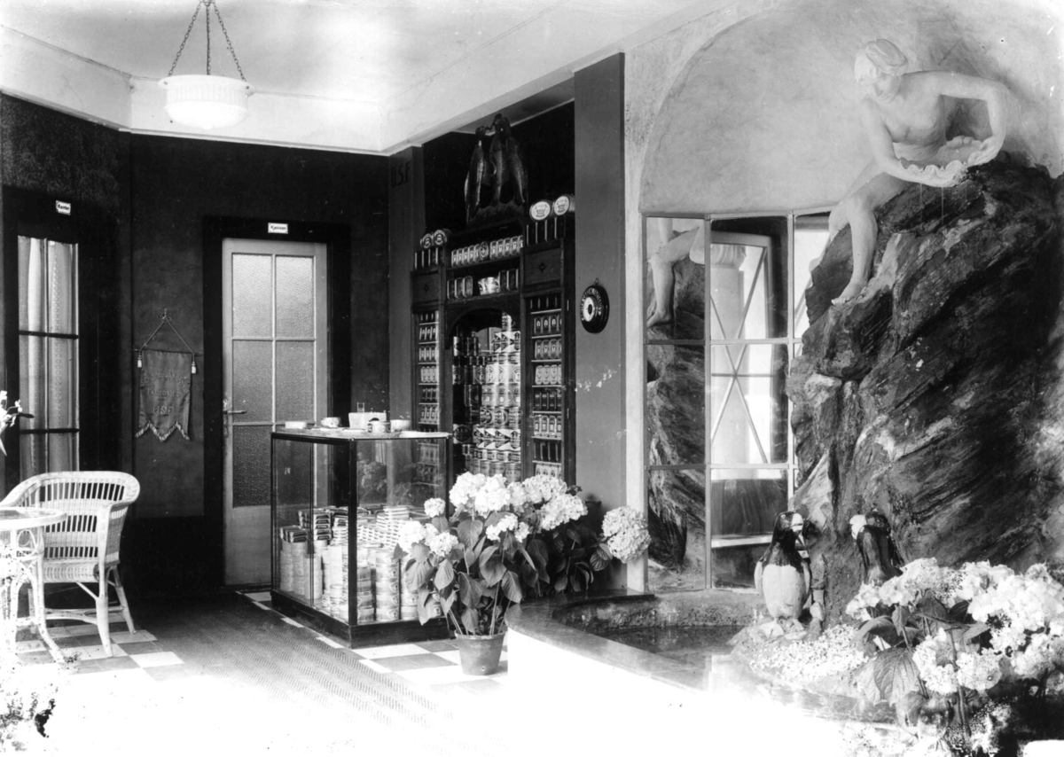 Hermann Christian Neupert, Jubileumsutstillingen på Frogner, Oslo, 1914. Interiør paviljong. Skulptur av kvinne. To lundefugler foran denne. Blomster på hver side av skulpturen. Reklameprodukter