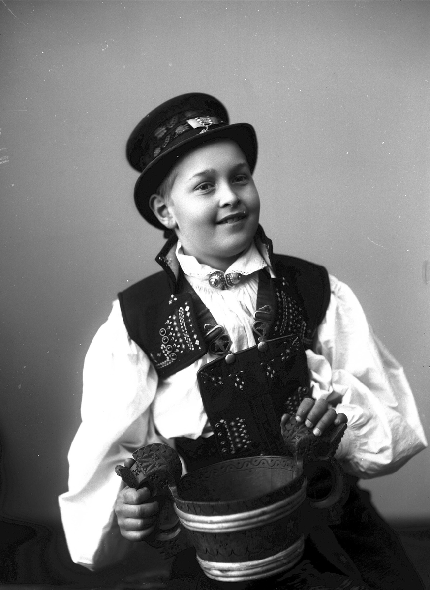 Solveig Lund, 1/2 portrett av gutt i drakt og hatt, tatt i fotografens studio. Han holder en staup påfanget. Nøytral bakgrunn.