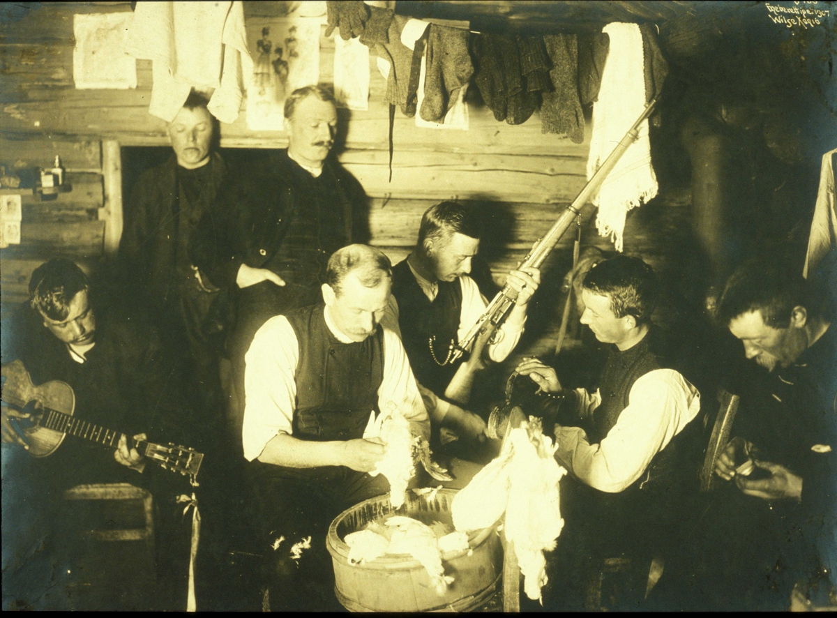 Anders Beer Wilse, Påsketur til Ellevestølene, Filefjeld ved Helevann 1907. NF.W 06468. Interiør med klær til tørk under taket. 7 menn utøver forskjellige sysler, blandt annet gitarspill, sjekking av gevær og plukking av ryper.