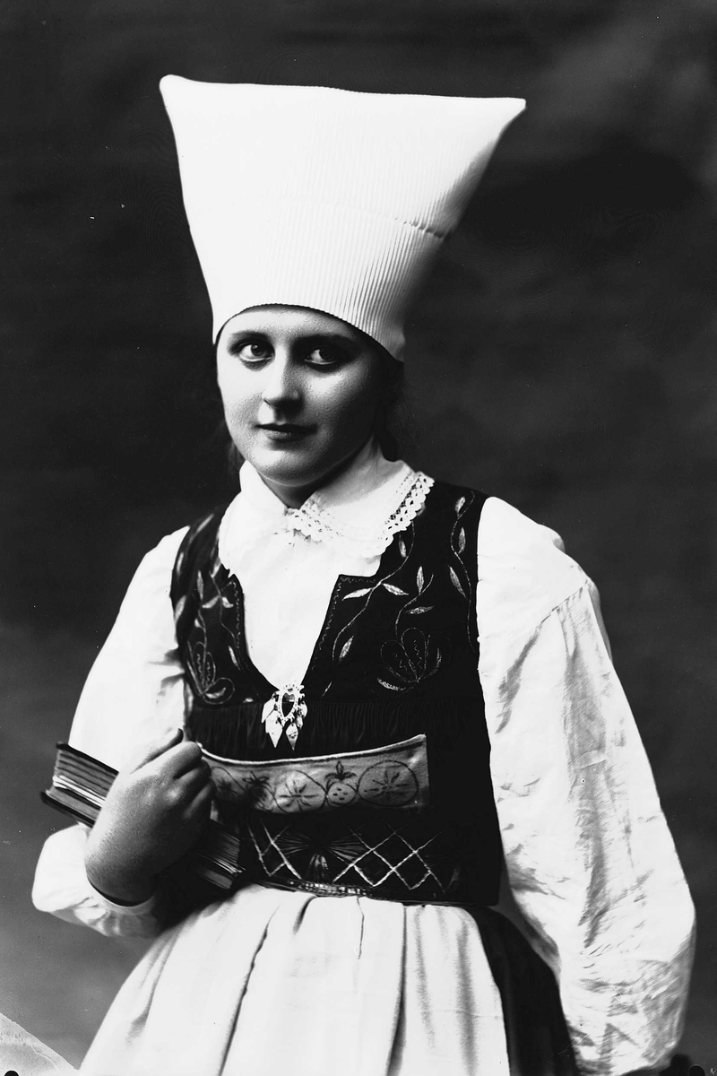 Solveig Lund, 1/2 dels studioportrett av kvinne i drakt og skaut. Hun holder en oppslått bok i venstre arm. Bakgrunnen er nøytral.