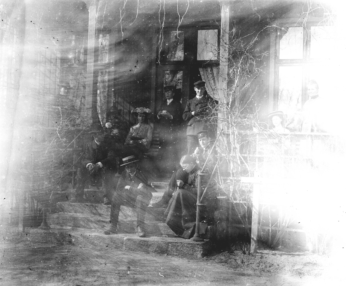 Juliane Fredrikke Langberg, Familiegruppe på veranda i kapellangården Bergenhus i Rakkestad, i anledning botanikkurs. Fotografens svoger, Olaf A. Holm, var res. kap. i Rakkestad 1884-97. Øverst t.v. står lærer Haaken Gran, g.m. prestedatter Margrethe, f. Holm, nederst t.h. Hennes bror Olaf Holm står bak i midten med kaffekopp…