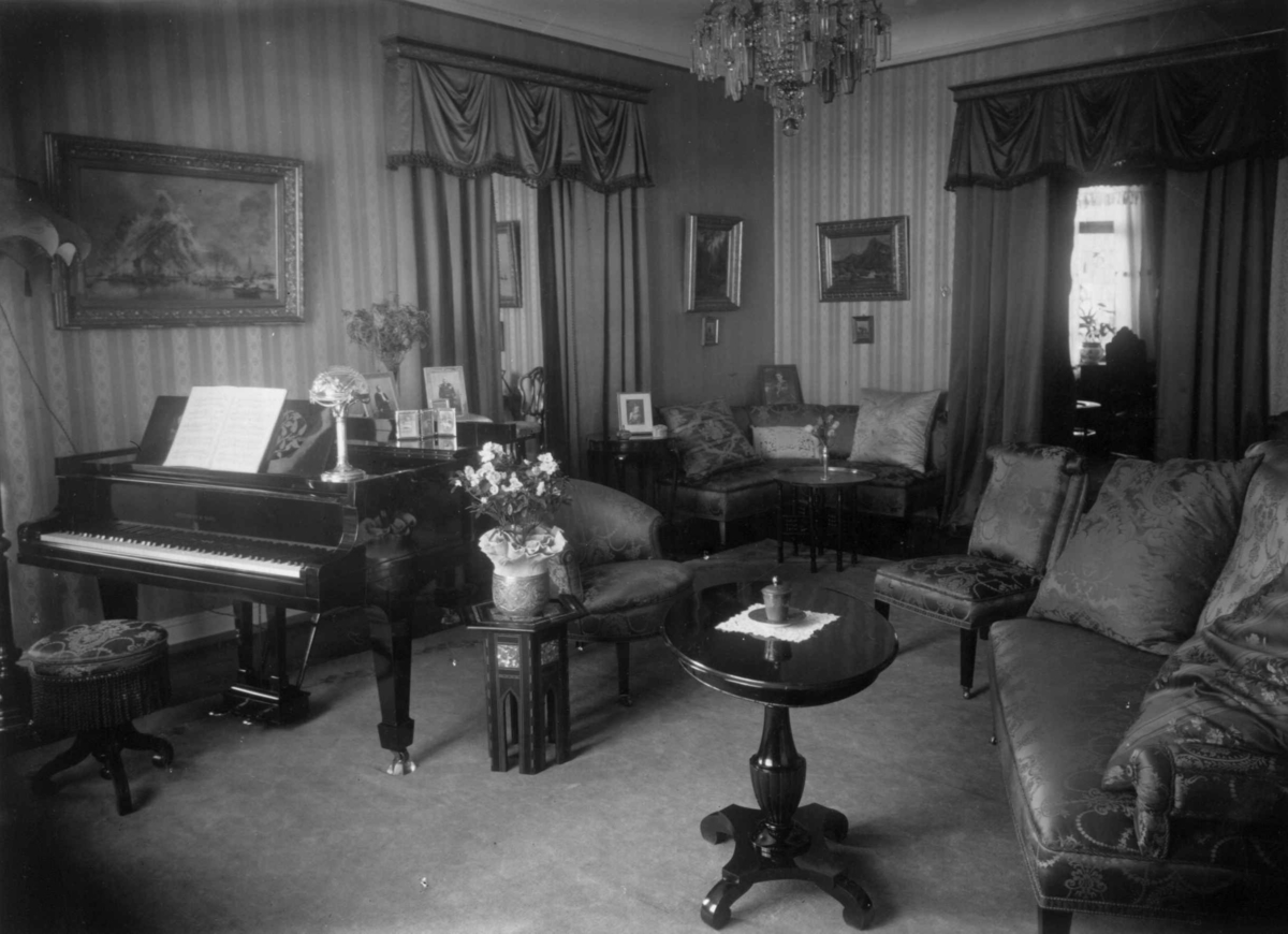 Hermann Christian Neupert, Trapp-Meyer. Stueinteriør 1925. Sofa med stoler og bord. Flygel. Malerier på veggene. Lysekrone.