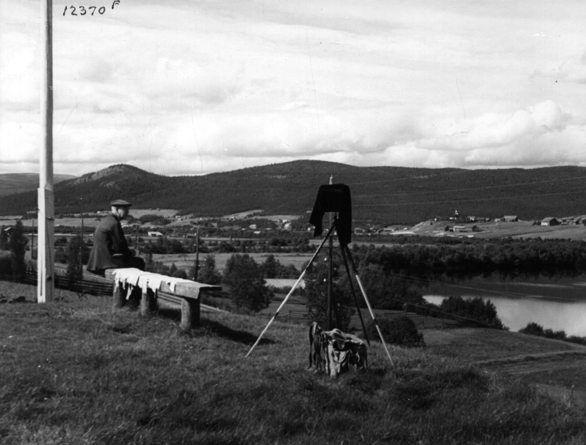 Anders Beer Wilse, Oversiktsbilde over Tynset, Hedmark, med Glomma, tatt fra benken på småbruket Hugubakken. I forgrunnen oppstilt fotoapparat. Fra Wilses protokoll vedr. personen på benken: "Högslund mediterer og korrigerer".