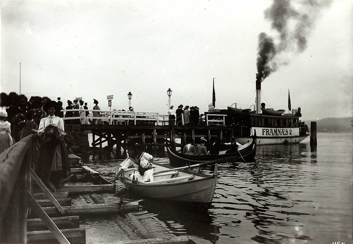 Hermann Christian Neupert, Oslo, Bygdøy 1908. Dronnningen restaurant. Mennesker som står i kø til ferja Framnæs 2 på brygga ved Dronningen. Kvinner i lange kjoler og med hatter. Robåter på vannet.