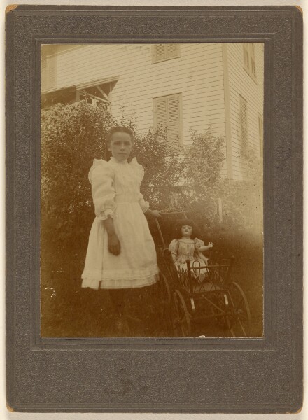 Unknown, [Unidentified little girl in white dress standing next to a baby carriage with a doll inside]
