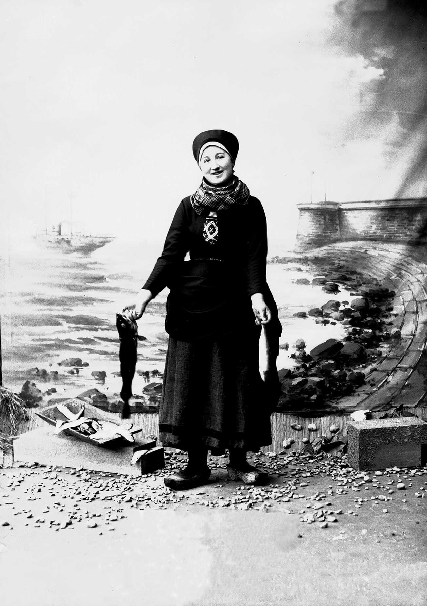 Solveig Lund, Studioportrett av kvinne i drakt med lue på hodet. Til høyre for henne ligger et trau med fisk. Hun holder en fisk i hver hånd. Bakgrunnen er malt med sjø, himmel, steiner, dampbåt og pir.