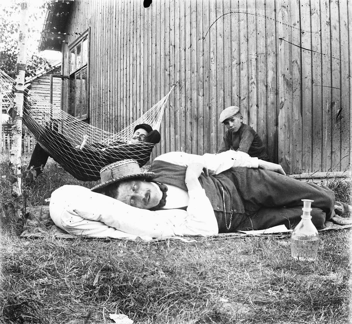 Ragnvald Andersen, To menn og en gutt slapper av ved et hus. Den ene i ei hengekøye. En karaffel til høyre.