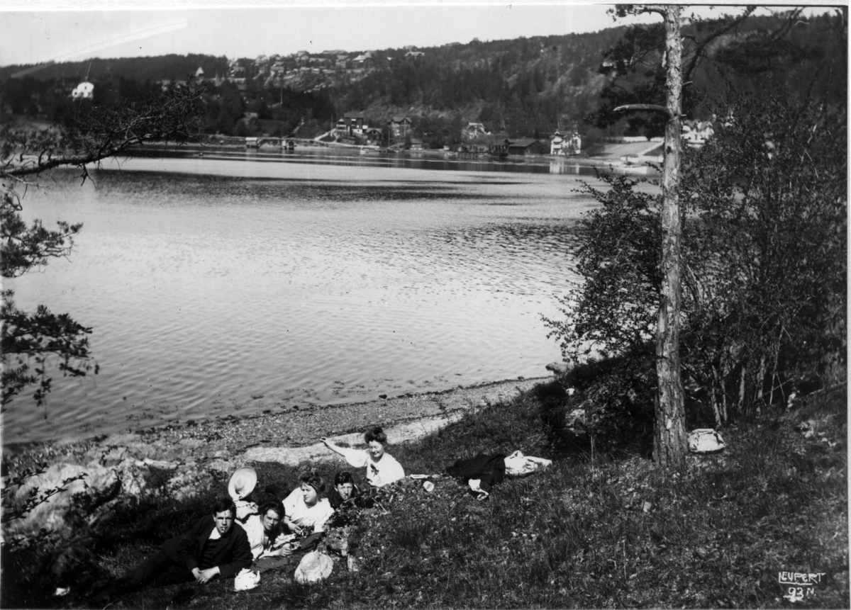 Hermann Christian Neupert, Ulvøya, Oslo 1908. Oversiktsbilde. Fjorden i forgrunnen. Gruppe med mennesker sitter på stranda. Bildet tatt mot fastlandet. Bebyggelse i bakgrunnen.
