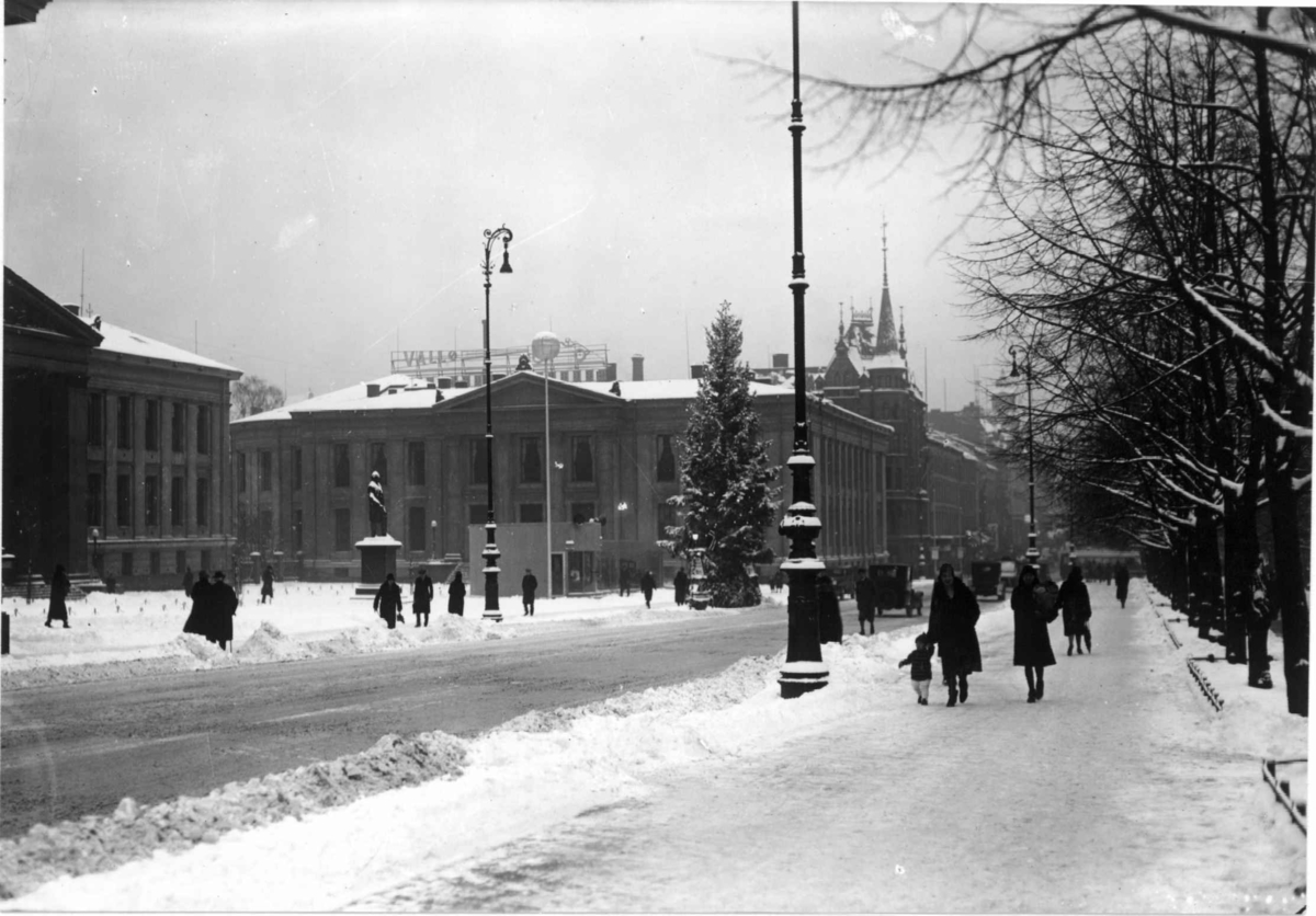 Hermann Christian Neupert, Oslo. Juletreet på Universitetsplassen. 1930. Snø. Karl Johans gate i forgrunnen. Universitetsbygningene i bakgrunnen. Fotgjengere.