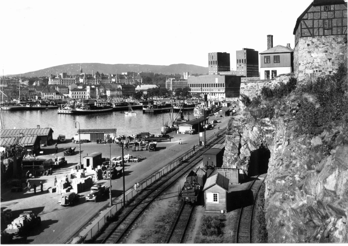 Hermann Christian Neupert, Oslo havn 1939. Utsikt over havna fra Akershus, med jernbanespor og vogner, biler, kaianlegg og Rådhuset. Victoria Terrasse og slottet i bakgrunnen. Fotgjengere.