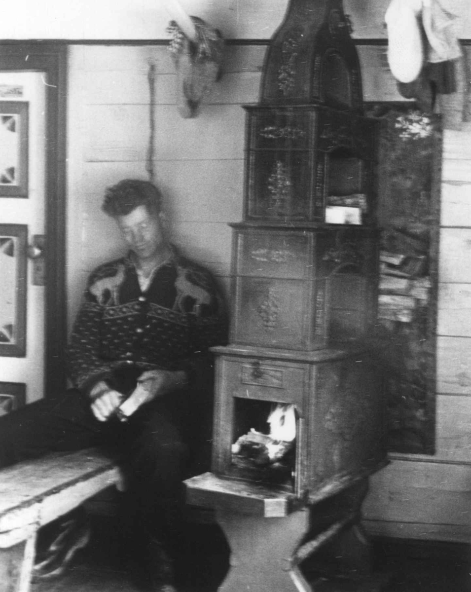 Anna Grostøl, Ola Ånonson sitter på en benk ved siden av vedovnen. Åseral, Vest-Agder 1943.