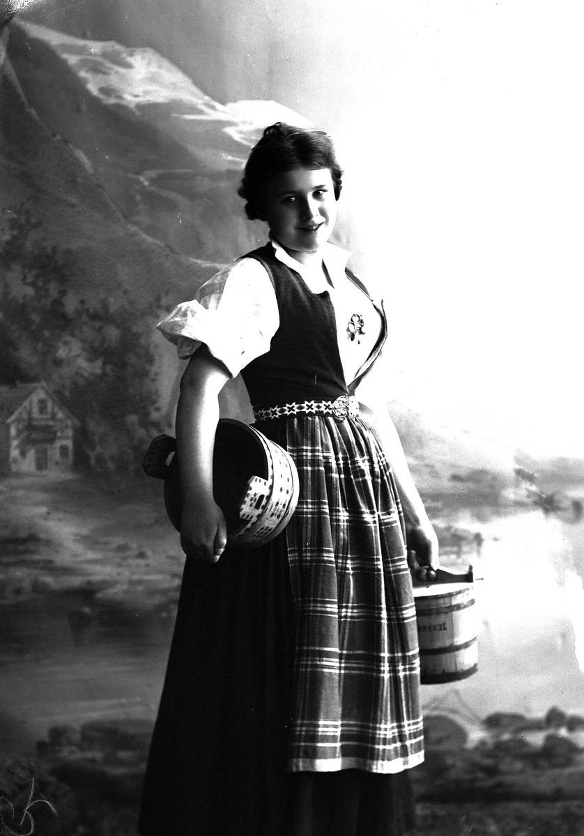 Solveig Lund, Studioportrett av kvinne i nasjonaldrakt som holder en tine under armen og en i hånden. Ser mot betrakteren. Bakgrunnen er malt med fjord, fjell og hus.