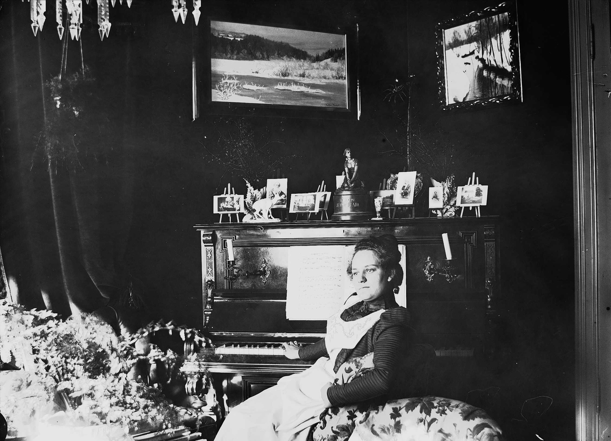 Axel Quinsgaard Wiborg, Kvinne i hvitt forkle sitter ved piano, Meltzers gate 9, Oslo, ant. 1899. Fotografier i ramme plassert på pianoet.