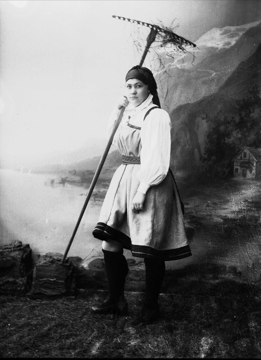 Solveig Lund, Studioportrett av kvinne i nasjonaldrakt som holder rive i den ene hånden. Hun er vendt mot venstre og ser ut på betrakteren. Bakgrunnen er malt med fjell, fjord og hus. På bakken er det gress.