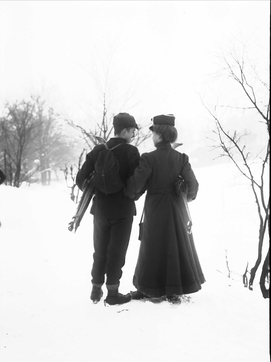 Solveig Lund, Mann og kvinne holder hverandre i armene i snødekt landskap. Fotografert bakfra - begge med ski under armen og mannen med sekk på ryggen.