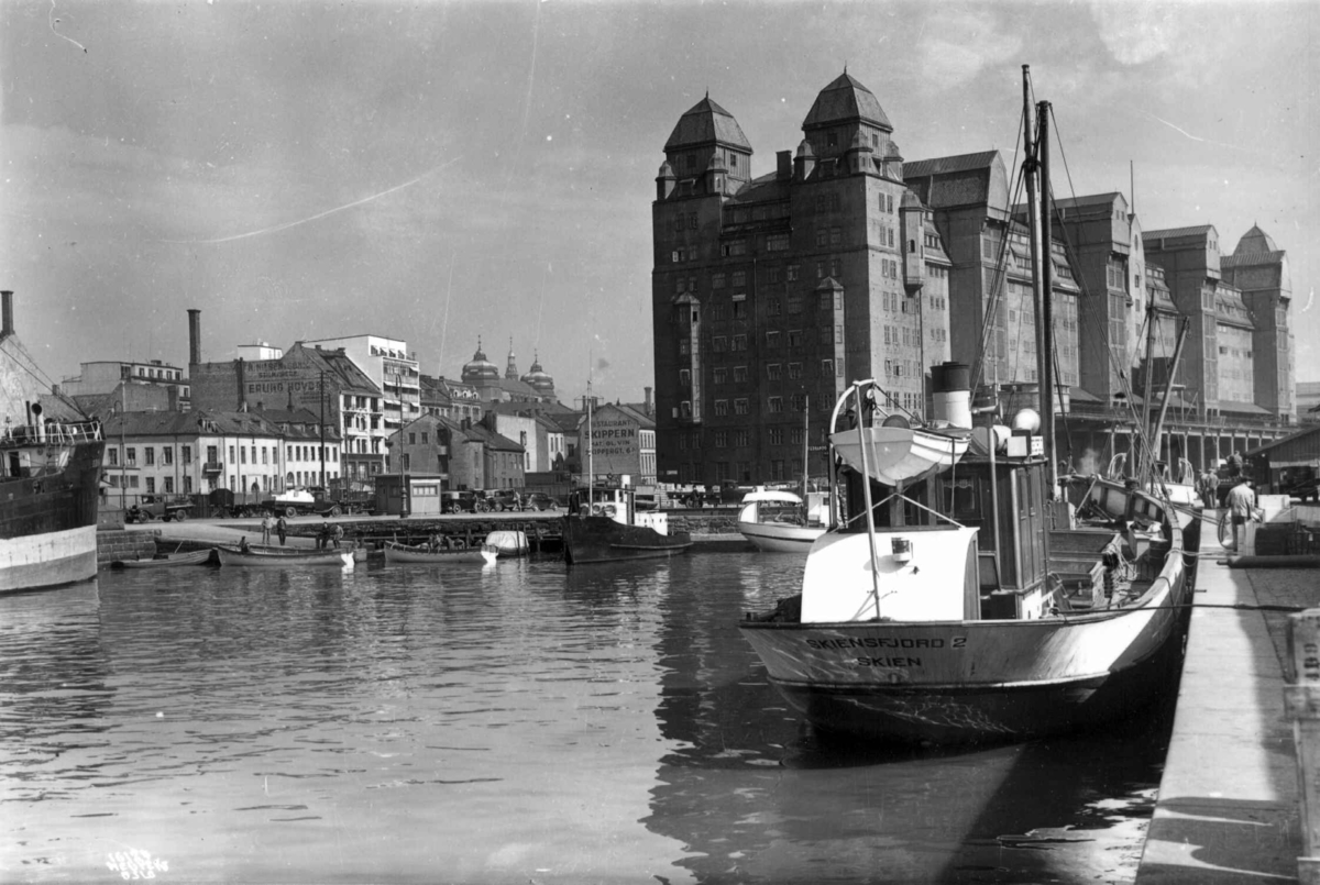 Hermann Christian Neupert, Havnelageret, Oslo 1937. Sett fra sjøen. Båter ved kai. Bebyggelse i bakgrunnen. Gate med biler og fotgjengere.