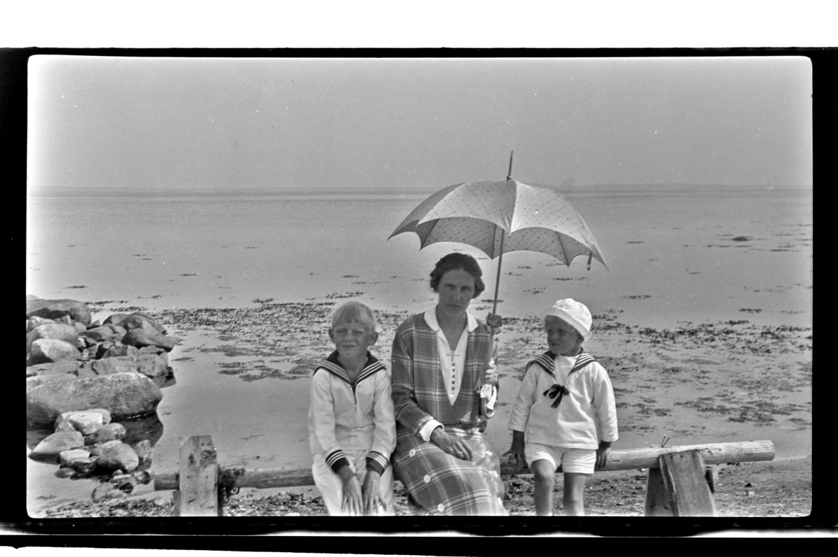 Rolf Sr. Sundt, Hilda Sundt, med parasoll, og barna Julius og Rolf Jr. på en benk ved ukjent strand. Fotografert ca. 1929.