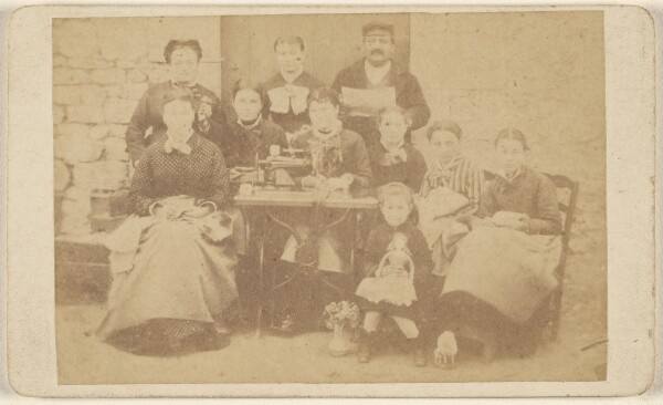 Unknown, [Group of ten men, women, and children sitting around a sewing machine]