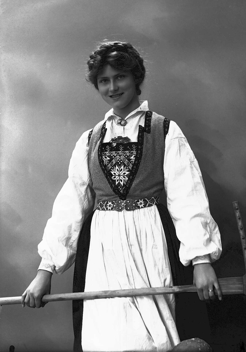 Solveig Lund, 3/4 dels studioportrett av kvinne i drakt, med oppsatt hår. Hun holder en rive foran seg. Bakgrunnen er nøytral.