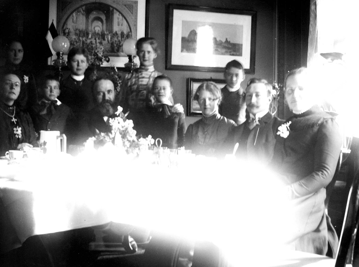 Juliane Fredrikke Langberg, Familiegruppe, hos sokneprest Olaf A. Holm, ant. på prestegården i Hurum (jfr. NF.15386-067). Gruppen sitter rundt dekket bord. Presten sitter i første rekke med skjegg. Helt t.v. fotografen. Som nr. 2 og 3 foran fra h. sitter Heinrich C. (Heini) Hudtwalcker og hans kone (fra 1903) Sigrid Holm, pre…