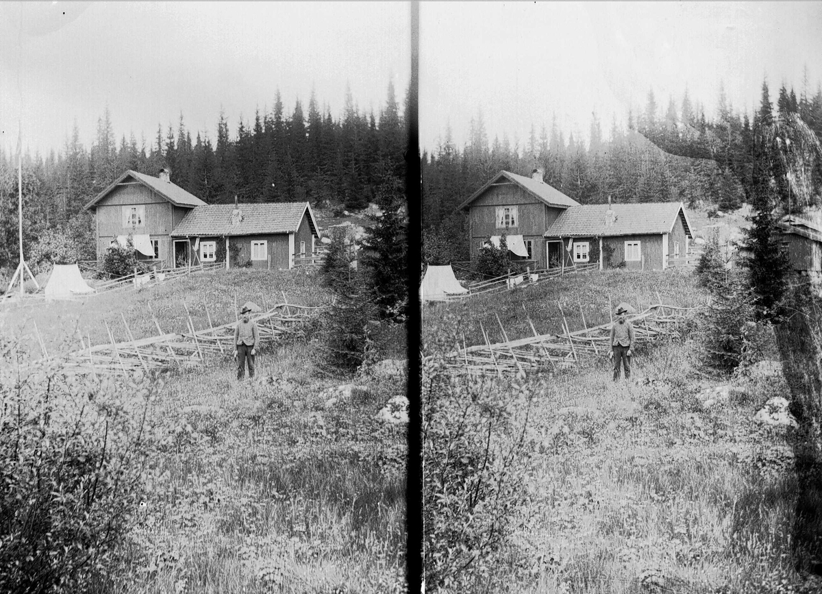 Axel Quinsgaard Wiborg, Mann med hatt (Gulbrand) står ved gjerde foran huset på Holoa seter, Hadeland, Jevnaker, Oppland, 1903.