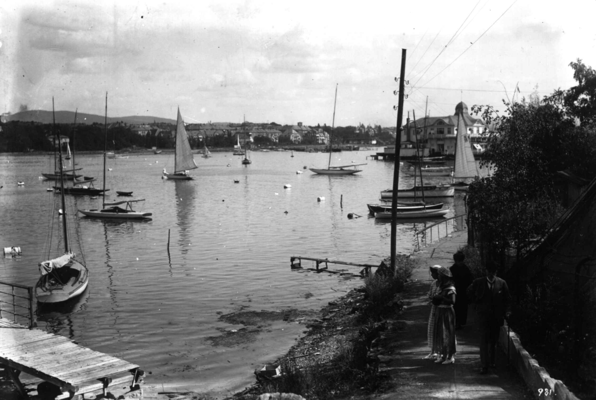 Hermann Christian Neupert, Oslo, Frognerkilen. To kvinner mot restaurant "Dronningen". Båter på fjorden. Brygger. bybebyggelse i bakgrunnen.