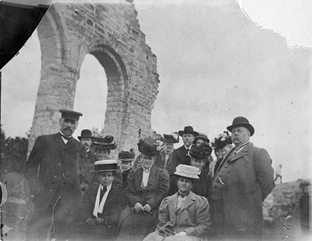 Anno Domkirkeodden, GRUPPE 14 PERSONER I DOMKIRKERUINEN VED SØYLEREKKA, YTTERST TIL HØYRE STÅR BAKERMESTER JOHS JOHANNESSEN OG FORAN HANS FRUE, HEDMARKSMUSEET OG DOMKIRKEODDEN