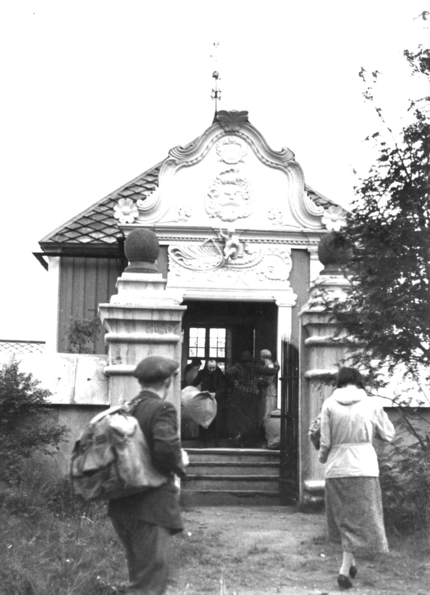 Anna Grostøl, Folk samlet for utdeling av ull i et lite hus med rik ornamentikk, Hjorts gravmæle. Røros 16. juli 1938.
