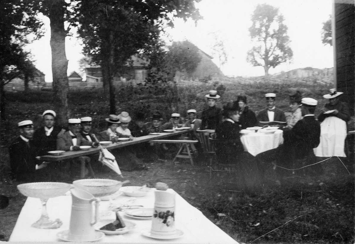 Okänd,, Kvinnor och män, troligen Harald Falk i studentmössa trea från höger med ansiktet mot kameran, sitter vid långbord, bord med skålar och tillbringare i förgrunden. Vid Grindstugan, Uppsala, omkring 1900