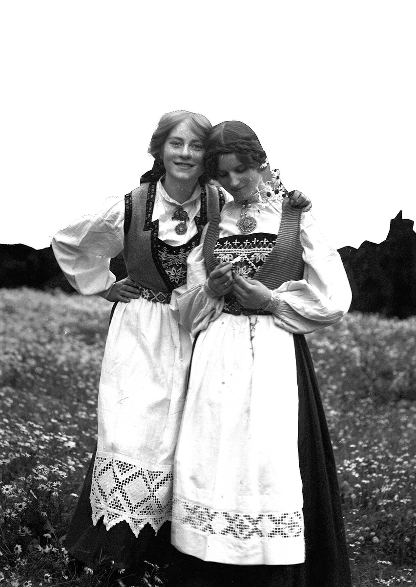 Solveig Lund, To kvinner i nasjonaldrakt står med armene rundt hverandre på blomstereng. Holder prestekrager i hendene.Fjell i bakgrunnen.
