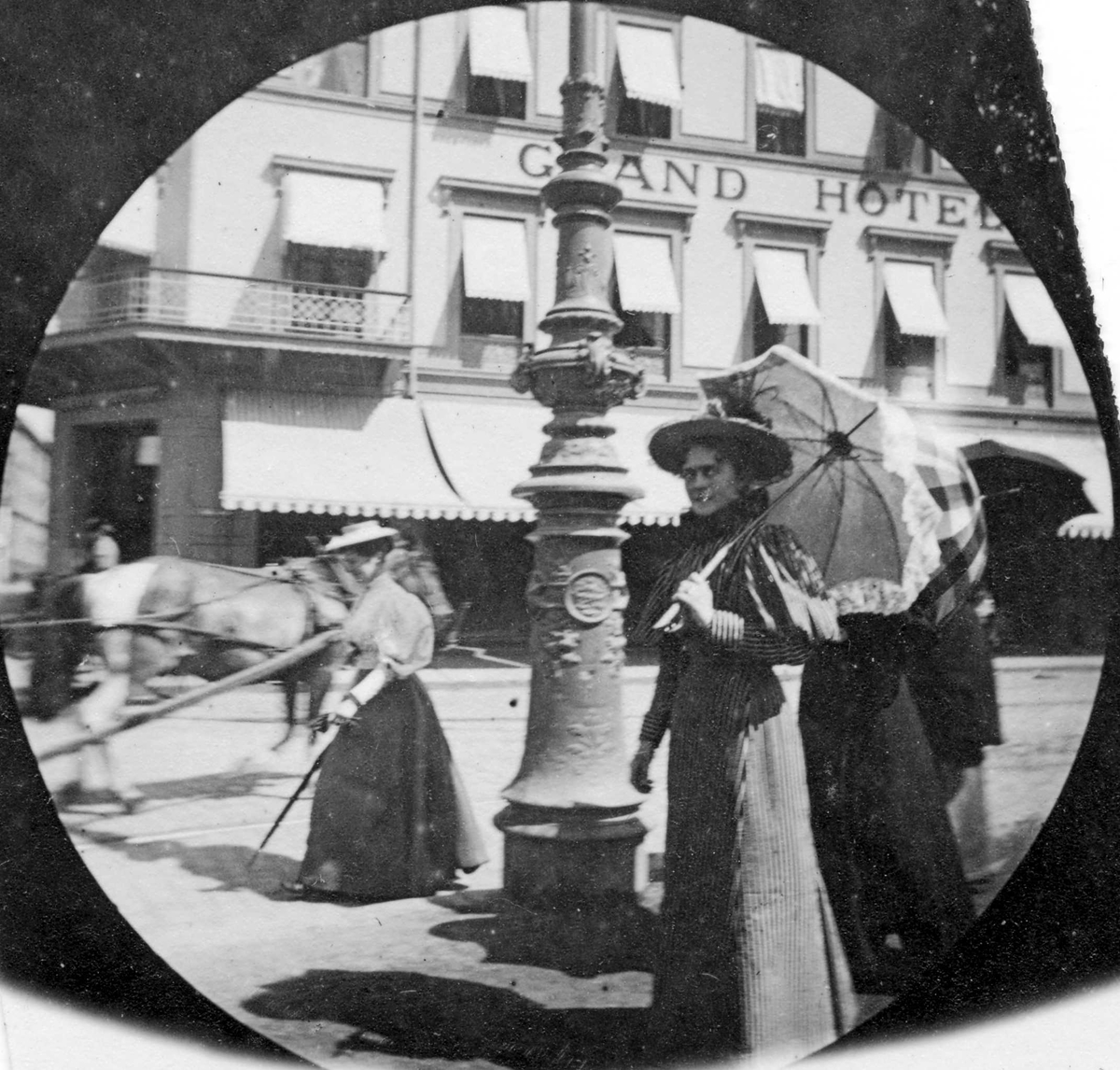 Fredrik Carl Mülertz Størmer, Frk. Røed spaserer på andre siden av gaten for Grand Hotell, Karl Johans gate, Oslo, med parasoll.