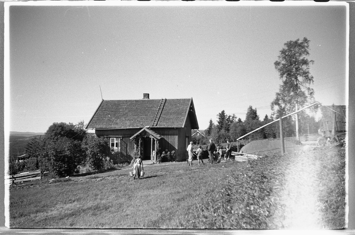 Håkon Prestkværn, Gruppe menn samlet foran et hus med vippebrønn. Kvinne bærer to sinkbøtter.