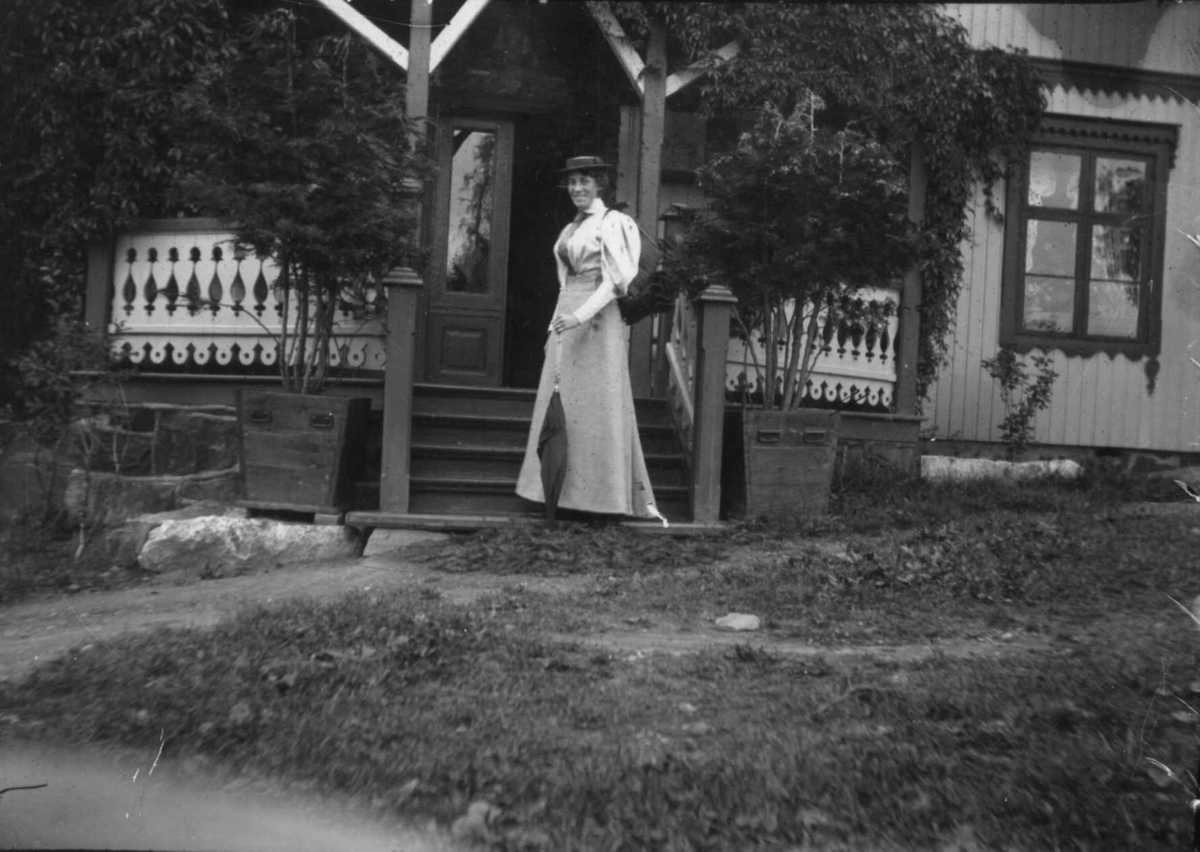 Hermann Christian Neupert, Drakt 1908-10. Kvinne stående i en trapp. I bakgrunnen hus med veranda.