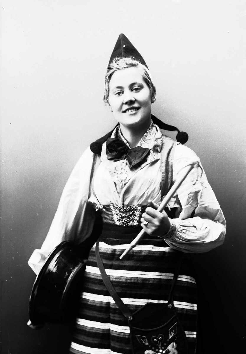 Solveig Lund, Kvinne i drakt og lue, som holder bolle under den ene armen og et redskap i den andre.