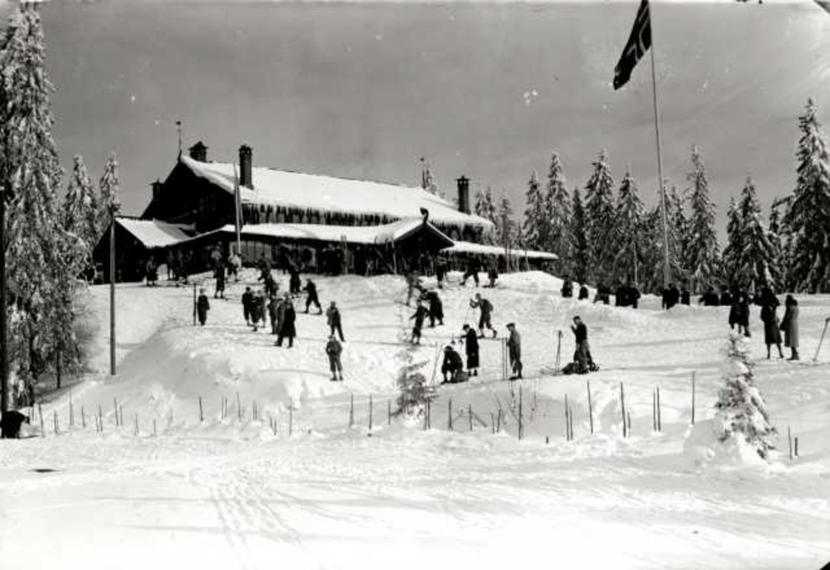 Hermann Christian Neupert, Frognerseteren Restaurant, Nordmarka, Oslo. Vintermotiv. Mennesker på ski foran resturanten. Flaggstang med flagg.