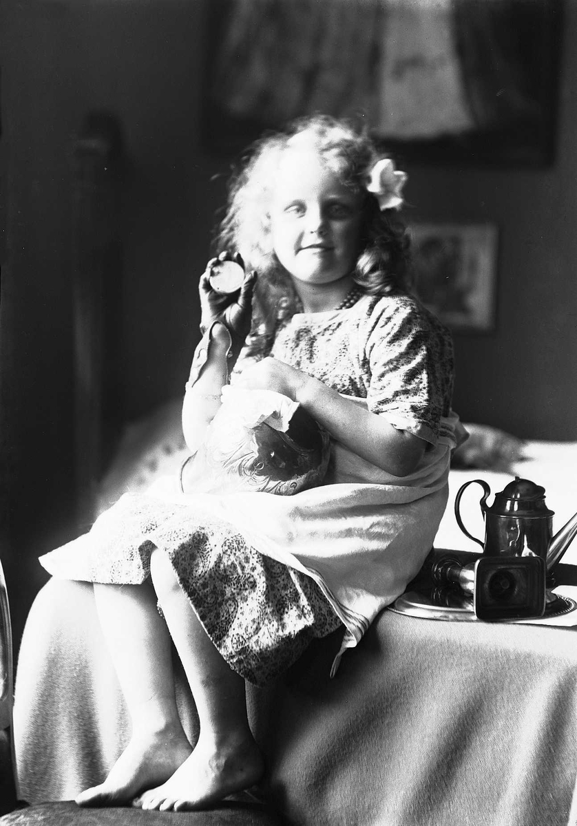 Solveig Lund, Pike sitter på bord med metallvase på fanget som hun pusser. På bordet ligger tallerken, lysestake og en kanne. Piken har perlehalskjede og blomst bak øret. Seng i bakgrunnen.