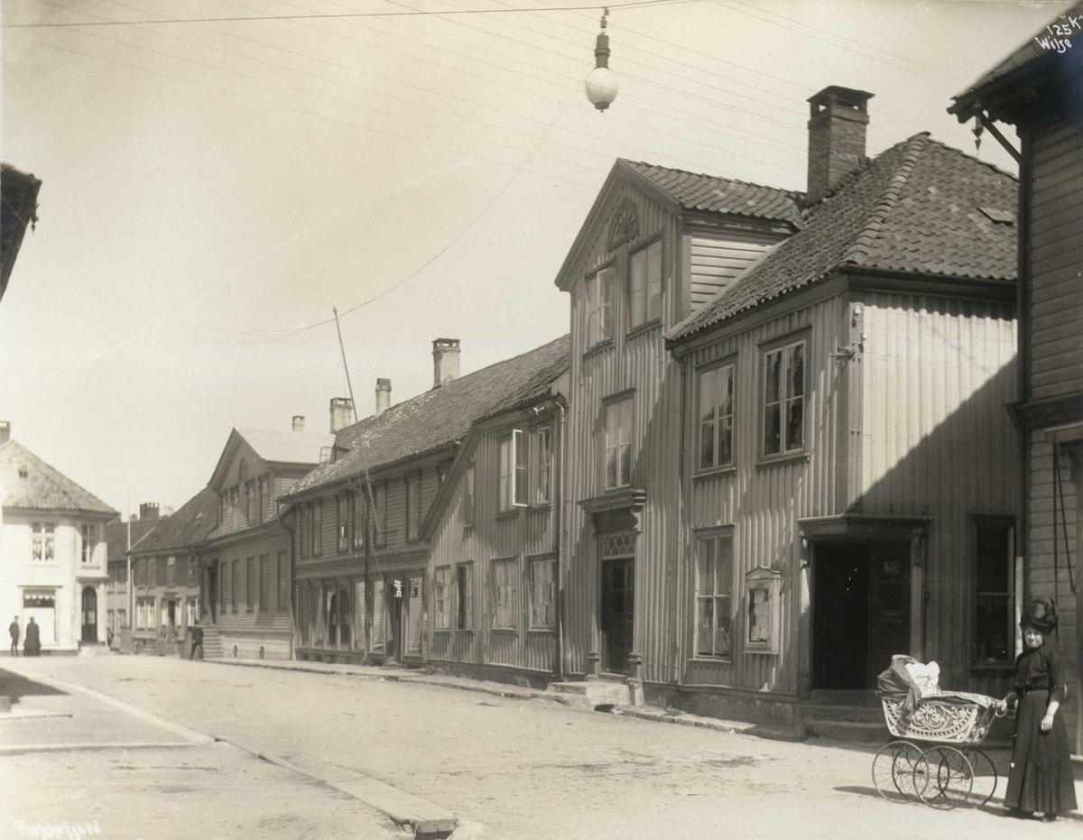 Anders Beer Wilse, Gateparti, Flekkefjord, Vest-Agder. Kvinne med barnevogn i forgrunnen. Fotografert 1912. Kaffistova til Mållaget i Flekkefjord skal ha vært etablert i dette huset i 1911.