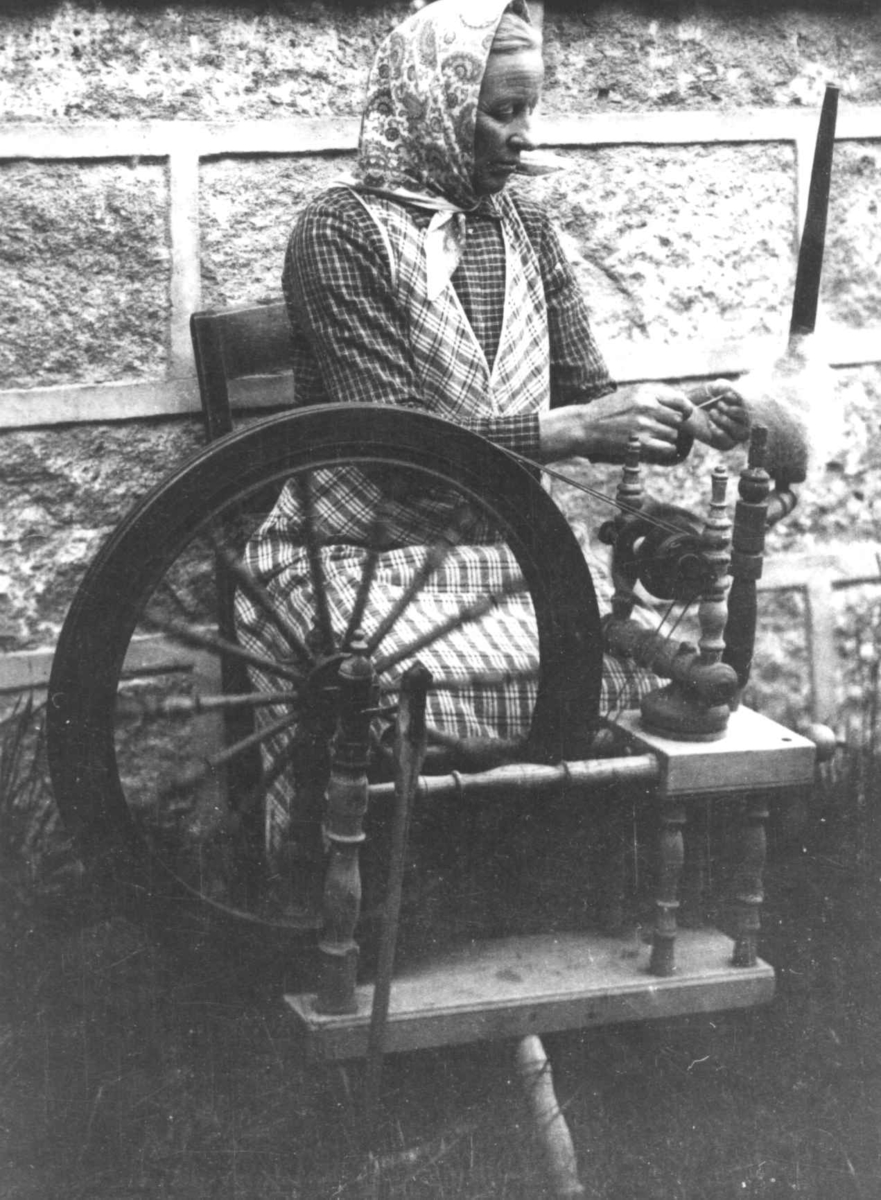 Anna Grostøl, En kvinne sitter ved rokken og spinner. Ukjent sted 1935.