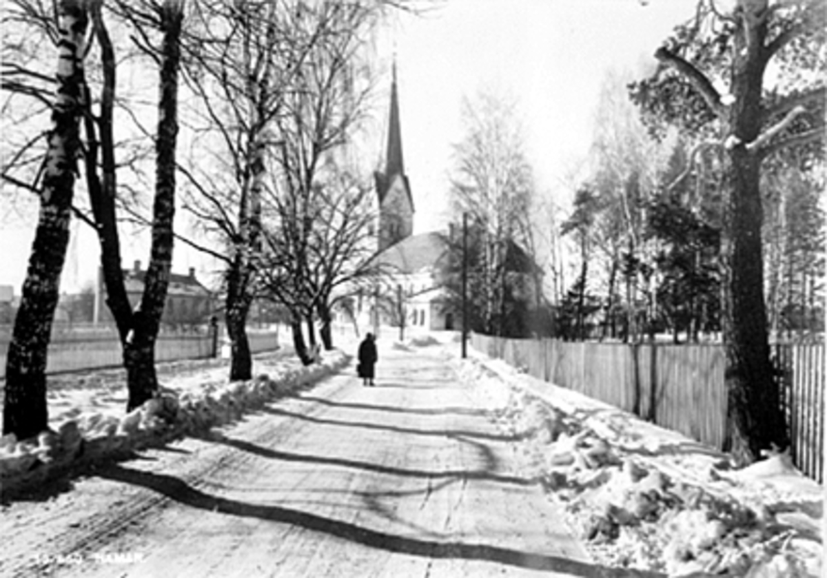 Normann Fotoatelier, Furnesvegen, Hamar. Vinter, Hamar Domkirke, Helga Lingjerde går nedover Furnesvegen. Postkort "Hamar"
