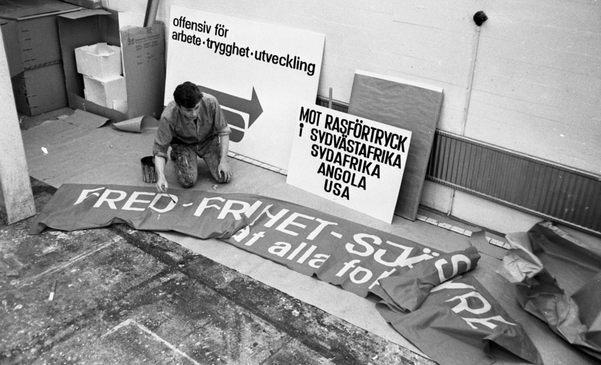 Örebro Kuriren, 1 maj förberedelser 26 april 1967 En man står på knäna och målar med en pensel på en banderoll inför 1 maj. Bakom honom står det skyltar lutande mot ett element.
