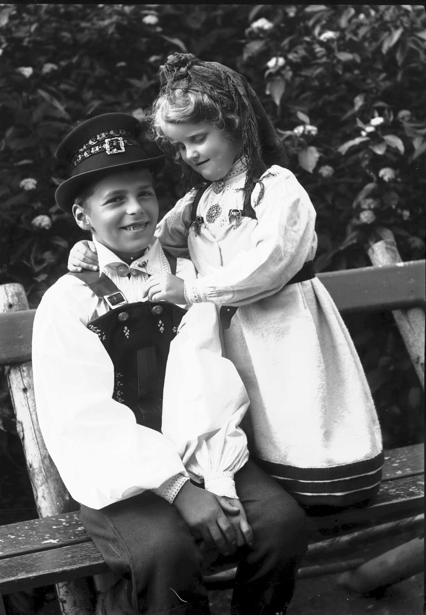 Solveig Lund, Gutt og pike i nasjonaldrakt. Sittende på benk, tre i bakgrunnen.