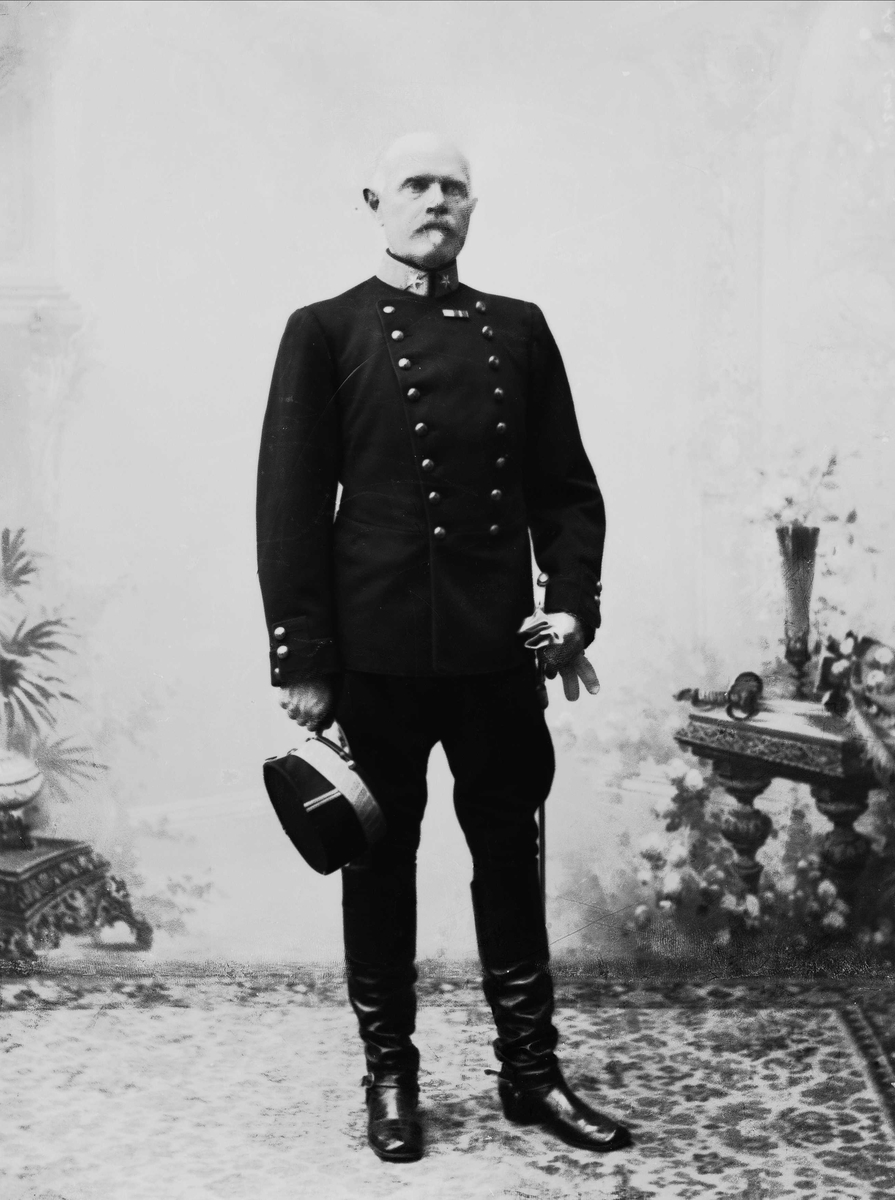 Solveig Lund, Studioportrett av eldre mann i uniform stående på teppe foran malt bakgrunn med blomster, bord og vaser. Holder lue i hånden.